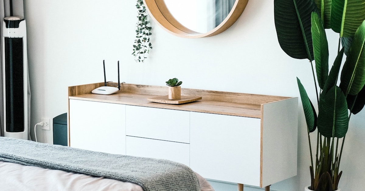 A bedroom with a low sideboard, a large round mirror on the wall above it and a tall plant beside