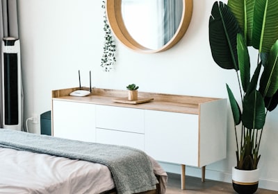 A bedroom with a low sideboard, a large round mirror on the wall above it and a tall plant beside