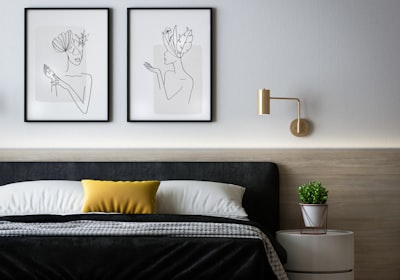 A bed with a dark headboard, two framed line drawings above it and a yellow cushion