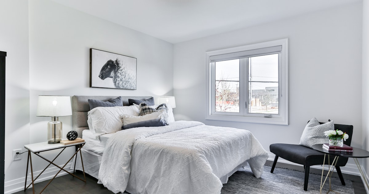 A white bed positioned under a bright window, with a bench at its foot