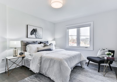 A white bed positioned under a bright window, with a bench at its foot