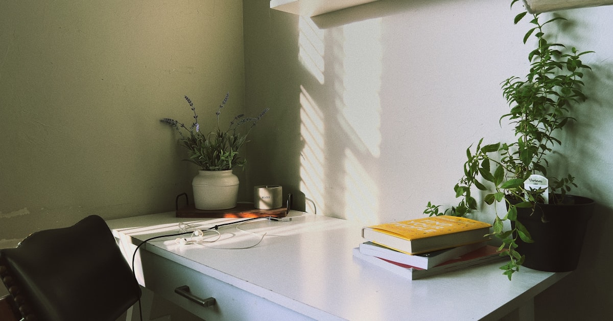 A small white desk against a green wall, holding a few books and two plants and nothing else