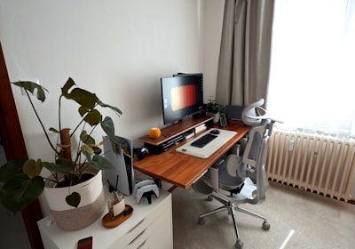 A desk with a monitor and keyboard, an office chair and a trailing plant beside the window
